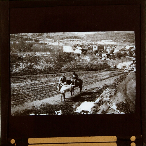 Two men on horseback in field by settlement