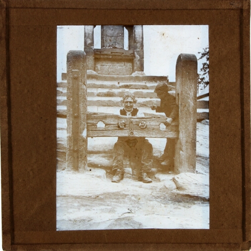 Man sitting in stocks, tickled with feather by young boy