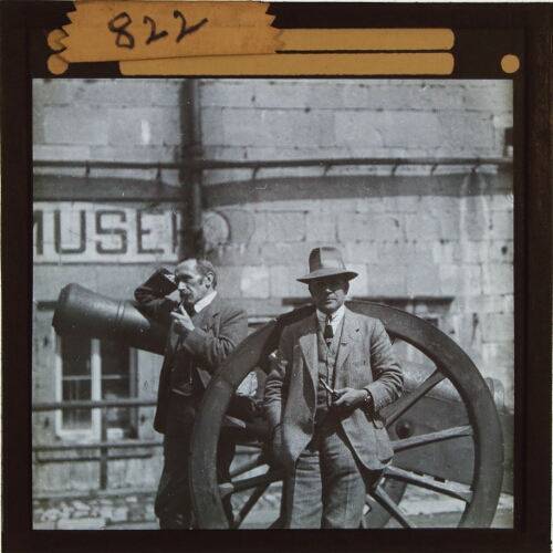 Two men standing by field gun outside museum in French town or village