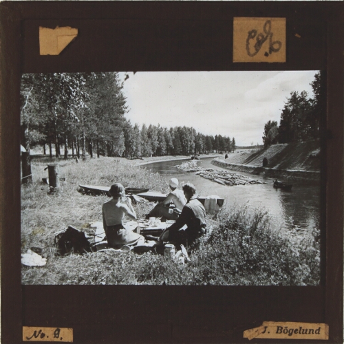 People picnicking by waterway with timber rafts passing