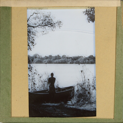Man standing by rowing boat at shore of lake or river