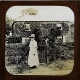 [Woman standing at garden gate of large house]