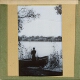 [Man standing by rowing boat at shore of lake or river]